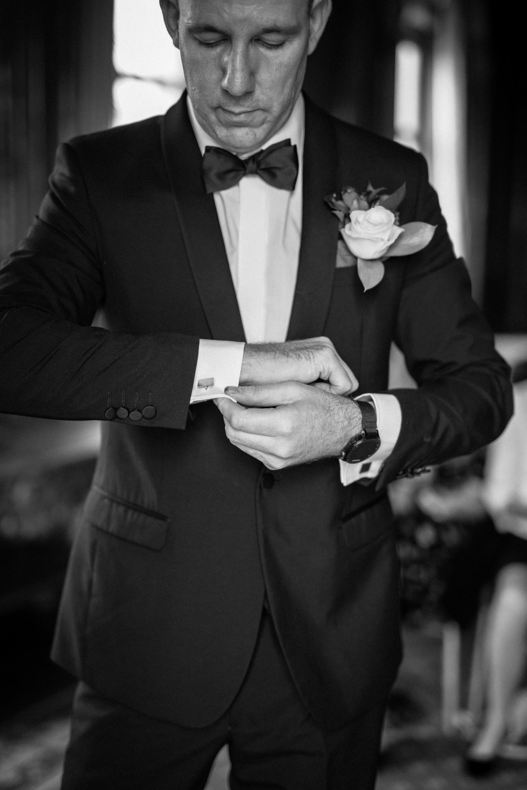 B&W. Cropped portrait of groom adjusting his cufflinks. Black suit and bow tie