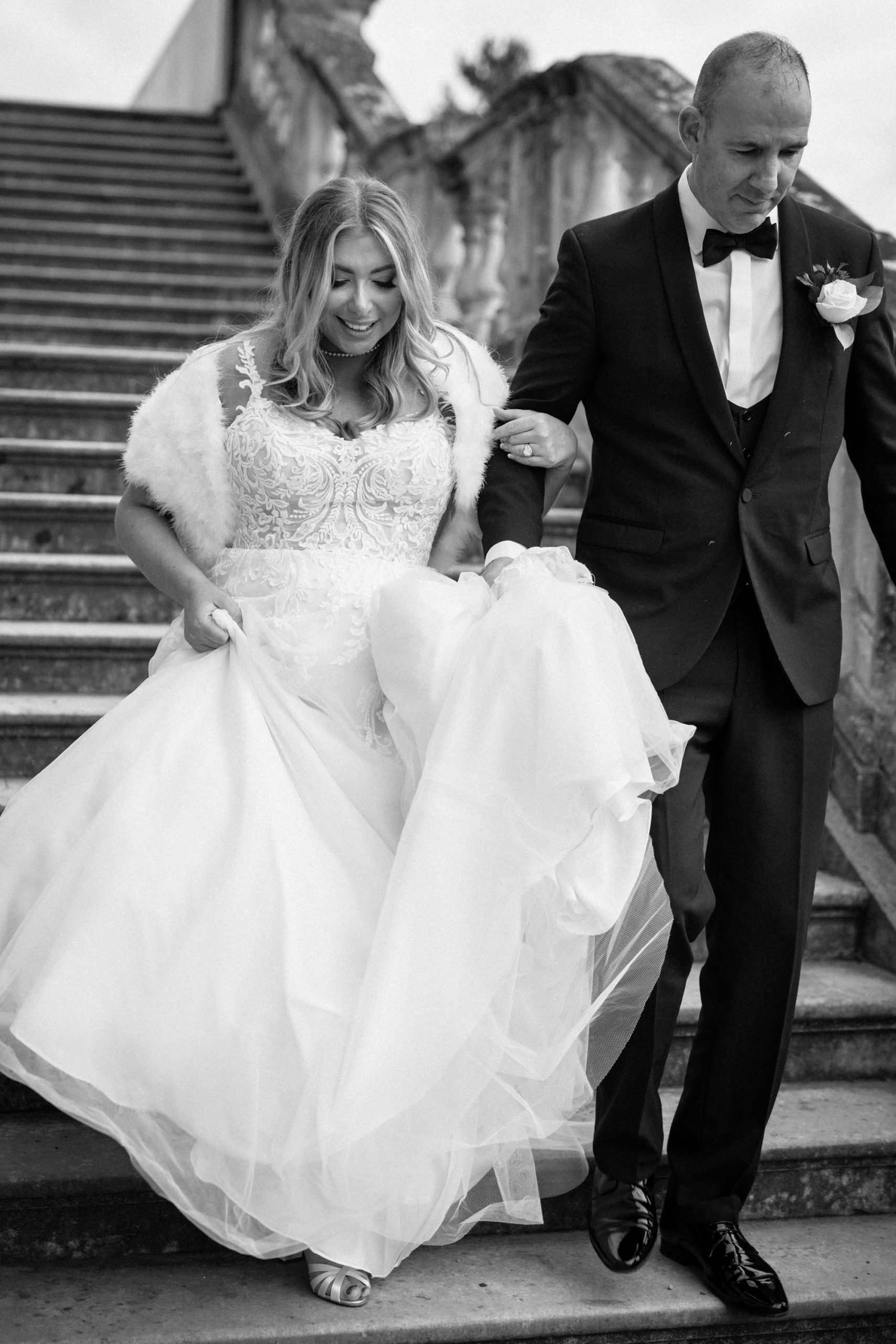 black and white image. Bride and groom walking down steps smiling. Groom wears black tie. Bride wears white wedding dress and white fur shrug.