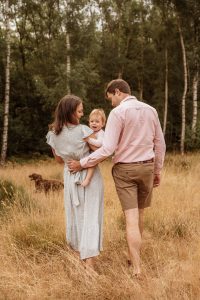 Mum and Dad walking away from camera in woods holding baby daughter. Dressed in Summer, pastel clothes.