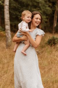 Mum in woods holding baby daughter. Dressed in Summer, pastel dress.