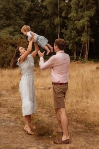 Mum and Dad in woods throwing baby daughter in the air. Dressed in Summer, pastel clothes.
