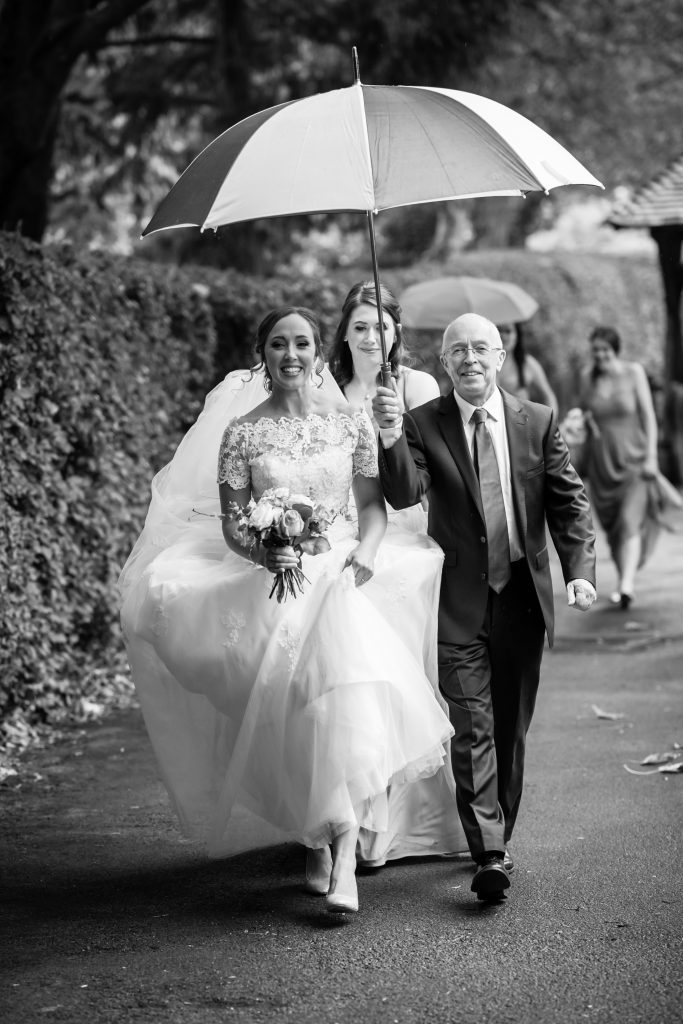 Bride walking to church with Dad next to her holding umbrella over her. Both smiling at camera. B&W photo