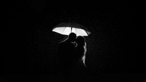 b&w photo of couple kissing in rain. Image is mostly totally black expect the back lit flash lighting up their silhouette and eth umbrella.