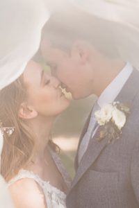 Bride and groom close up on face. The veil is over their heads and they are kissing.