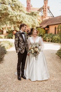 Groom in tux and black tie, bride in long sleeved wedding dress looking at camera, holding fresh flowers in green, cream and burnt orange.