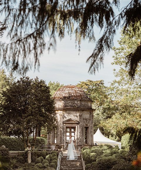 View of the chapel at The Larmer Tree wedding venue with foliage and trees in foreground. Bride stood on steps smiling.